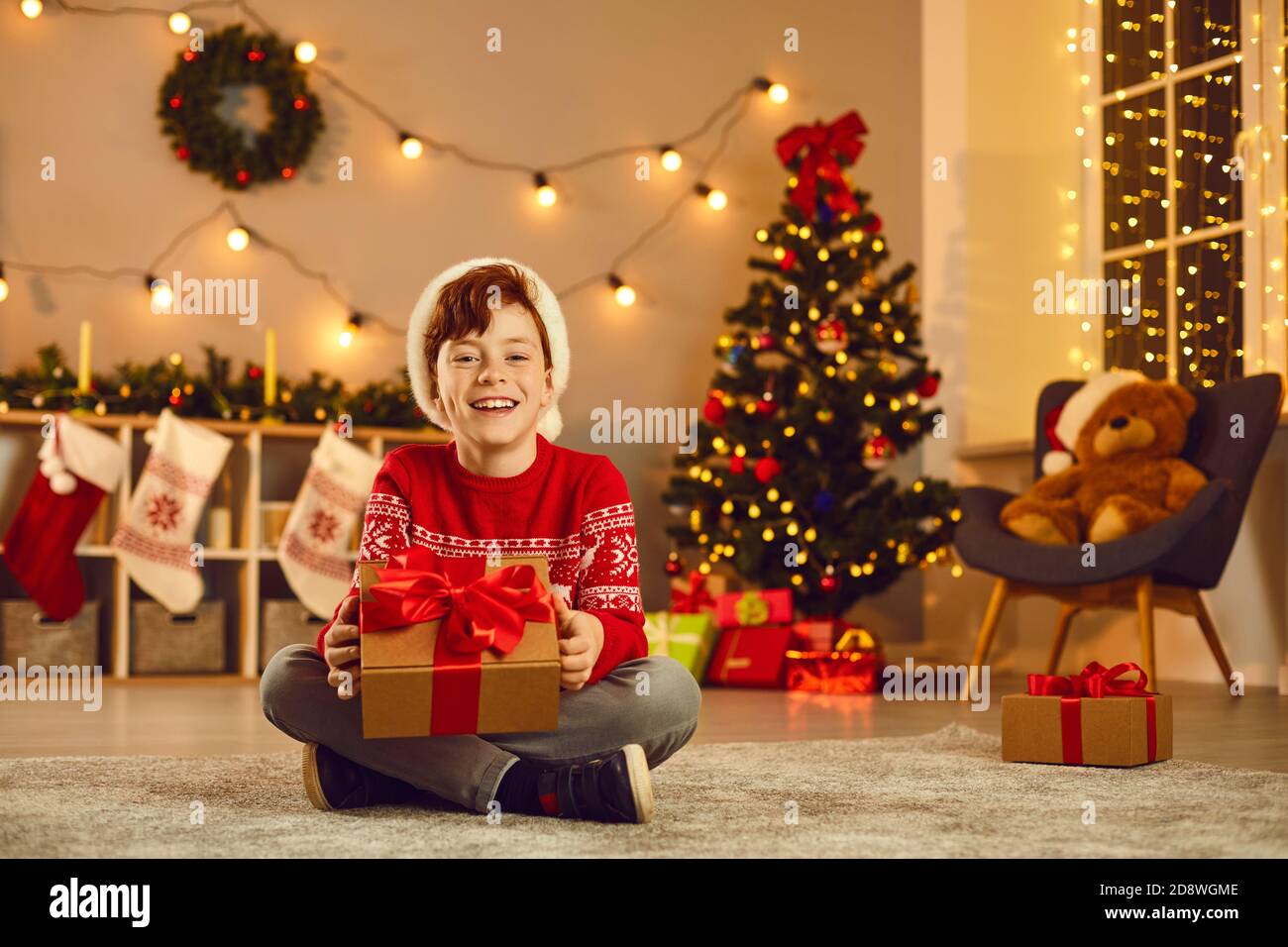 Niño sonriente sentado en el suelo y sosteniendo la caja de regalo Las manos durante la Navidad Foto de stock