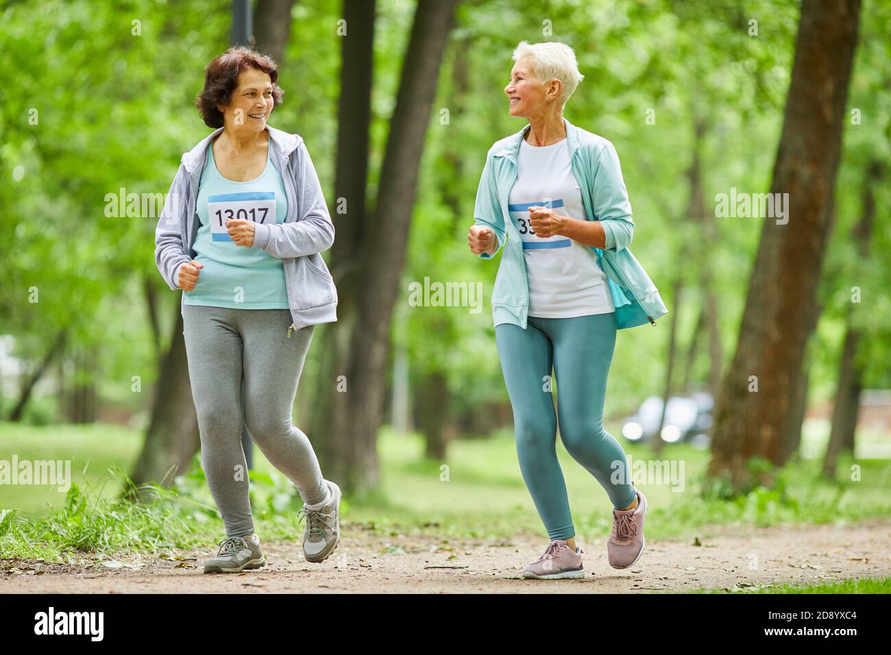 Tiro de cuerpo entero de dos mujeres mayores activas corriendo maratón carrera en el parque de la ciudad hablando entre sí Foto de stock