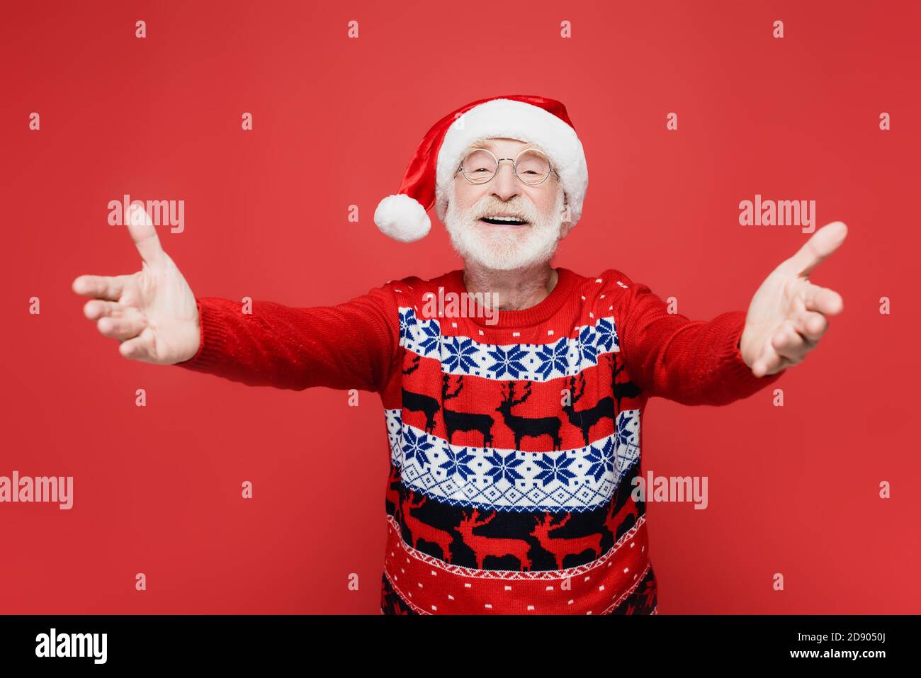 Hombre mayor sonriente en el sombrero de santa tirando de las manos en la cámara en primer plano borroso aislado en rojo Foto de stock