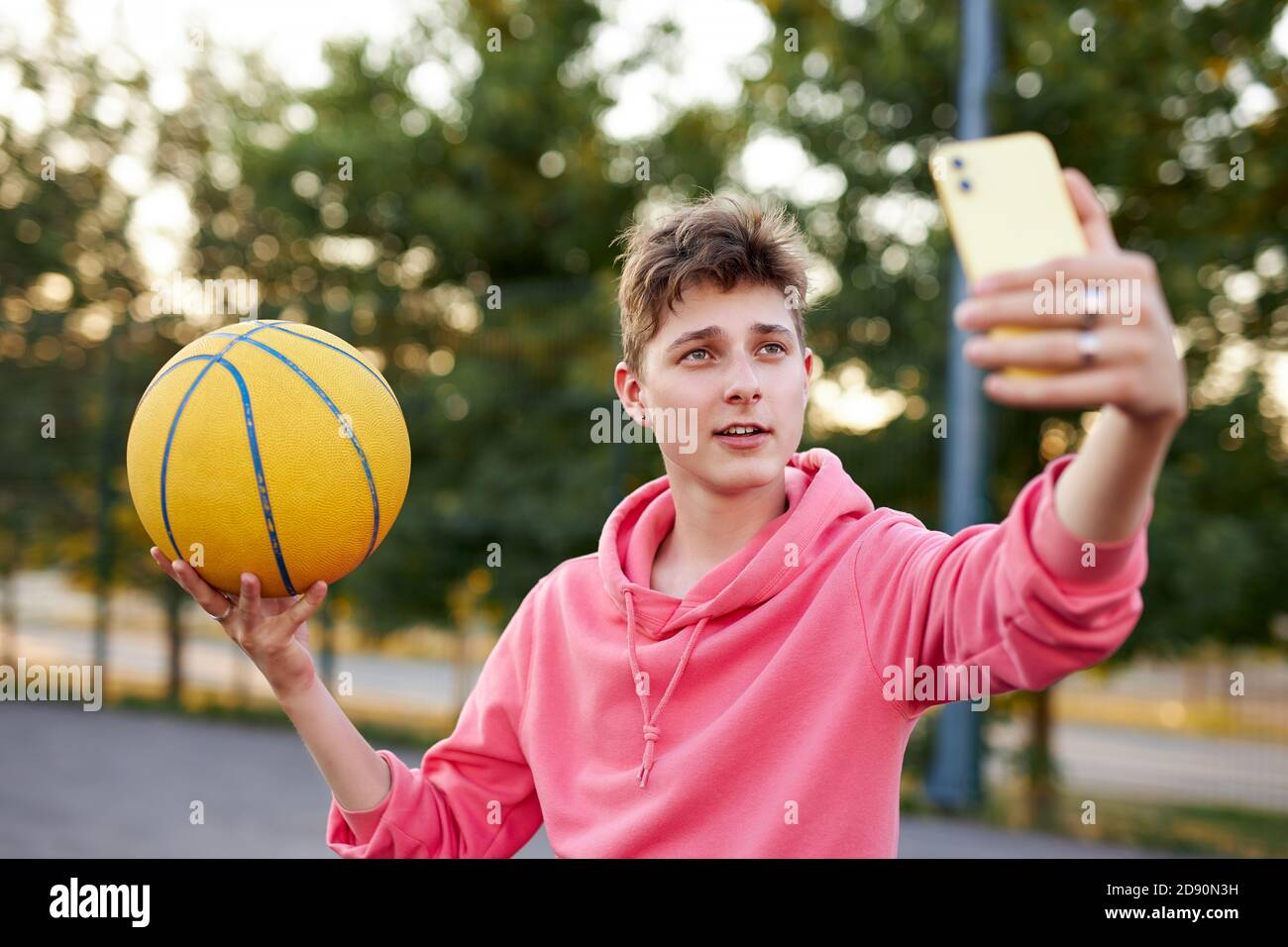 joven basketballer caucásico tomar una foto de sí mismo con pelota, chico atlético mirar smartphone, posando Foto de stock