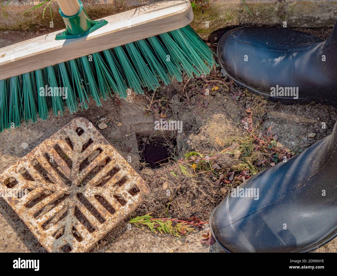 Una persona en wellington botas con una escoba de madera, con una pequeña cubierta de drenaje quitada, a punto de barrer los desechos acumulados alrededor de un agujero de remozo. Foto de stock