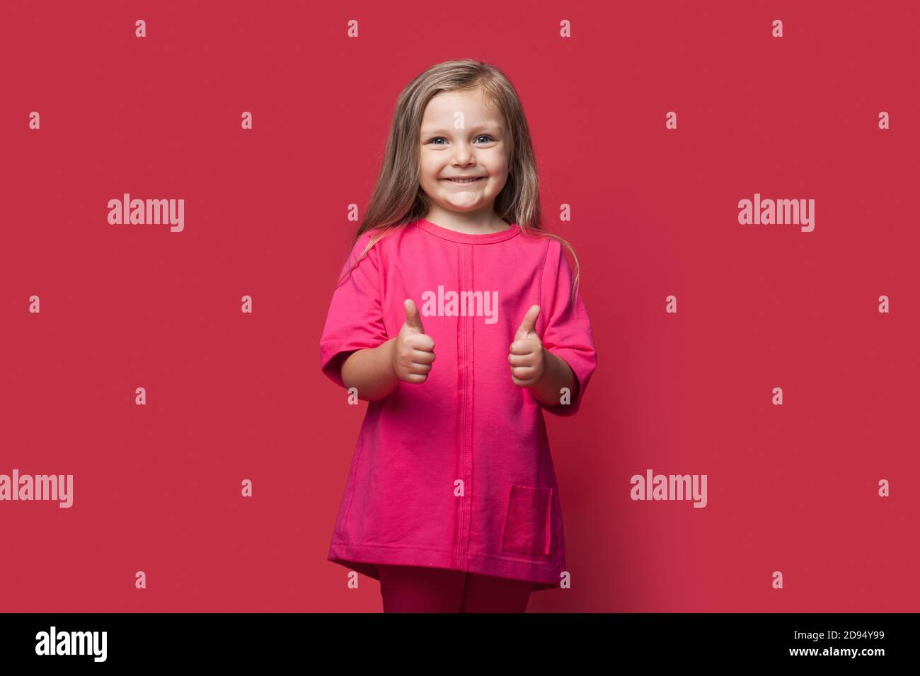Foto monocroma de una chica rubia gestando el signo similar en una pared roja del estudio sonriendo a la cámara Foto de stock
