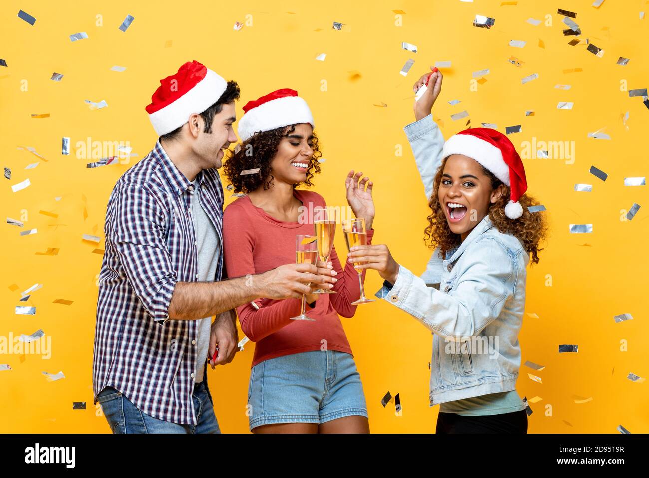 Grupo de amigos diversos bebiendo champán celebrando la Navidad en amarillo fondo de estudio de color con confeti Foto de stock