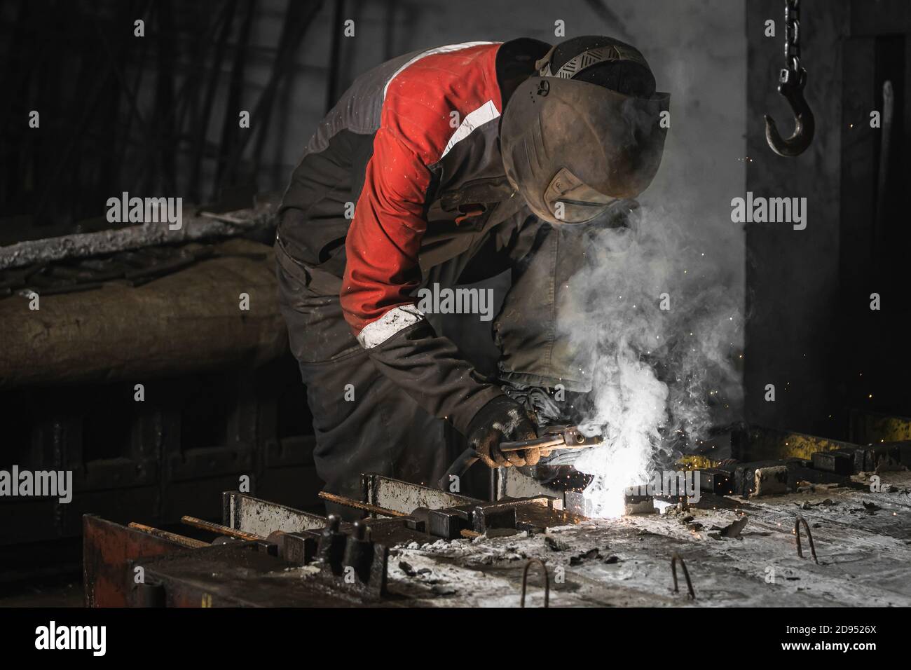 Cerca de un hombre fuerte es un soldador en uniforme, máscara de soldadura  y cuero de los soldadores, un producto de metal se suelda con la máquina de  soldadura en la obra