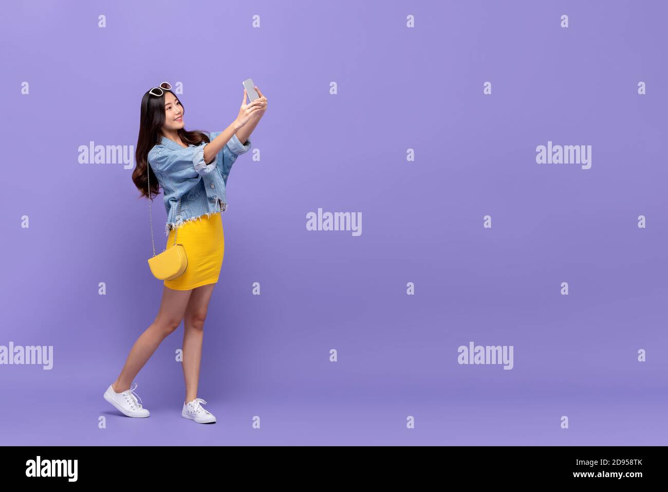 Retrato completo de una joven y sonriente mujer asiática que toma selfie con smartphone en el estudio aislado fondo púrpura con copia espacio Foto de stock