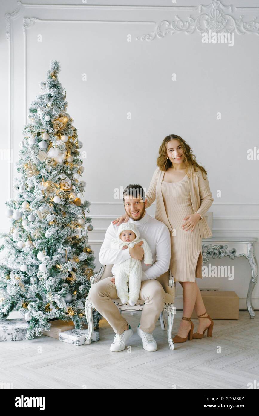 El concepto de una Navidad familiar. Una hermosa familia con ropa ligera  está sentada junto a un árbol de Navidad decorado. Feliz familia con un  bebé en una ch Fotografía de stock -