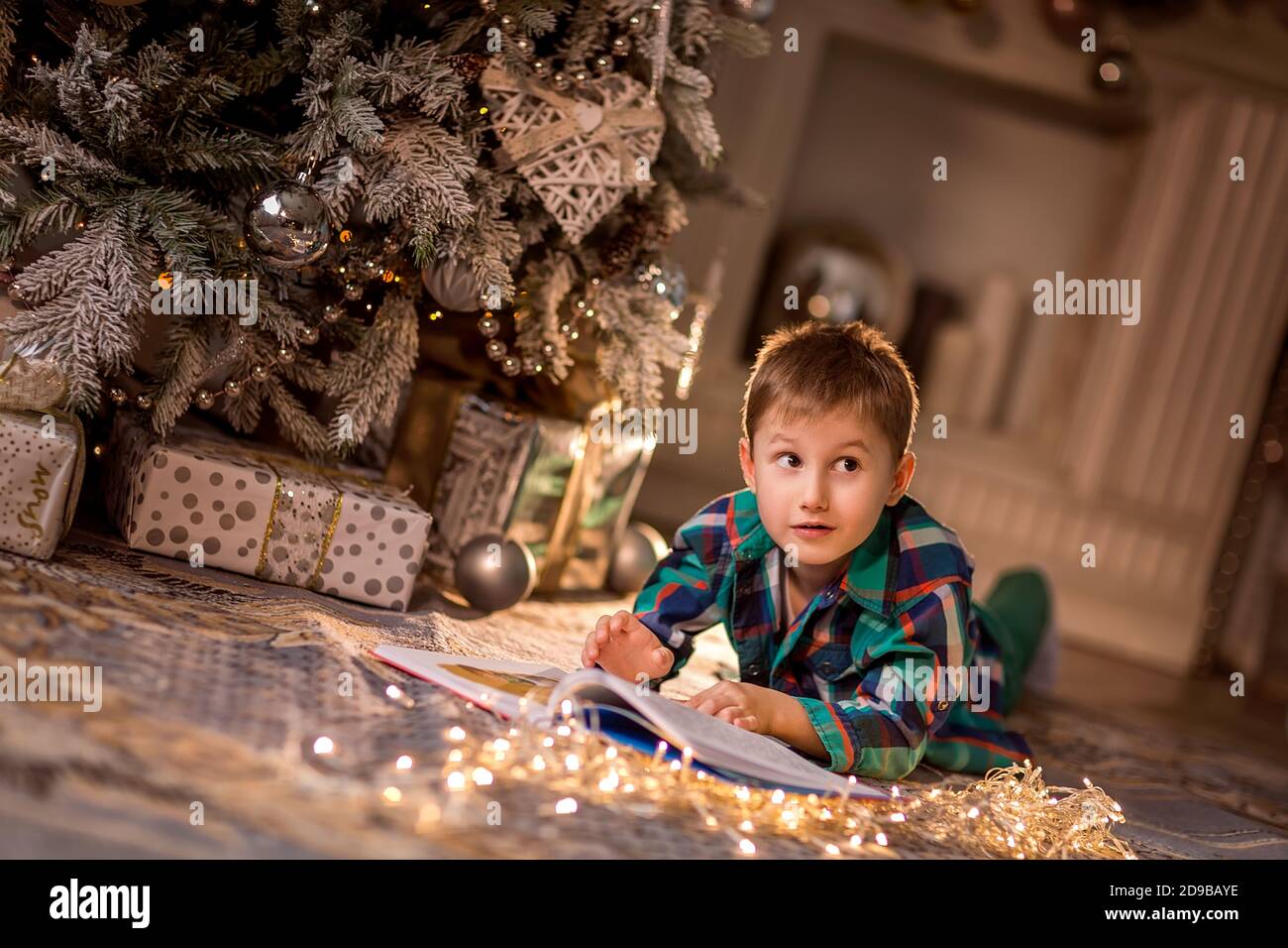 El niño está acostado en el suelo cerca de un árbol de Navidad decorado. Leer un libro con historias de Navidad. Acogedoras noches de invierno en casa. Concepto de Cristo Foto de stock
