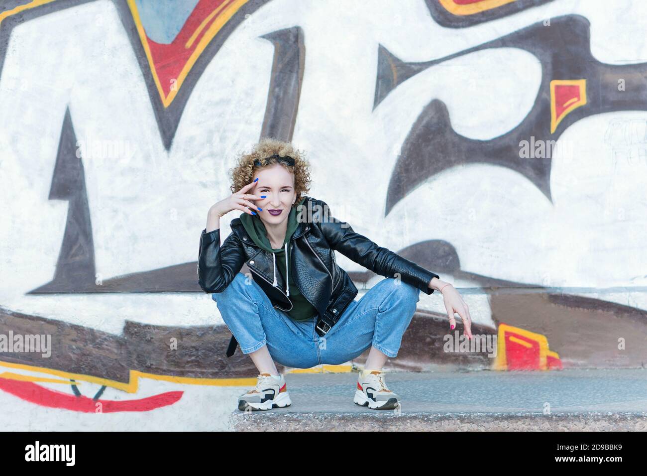 Retrato completo de una chica de cintura baja de moda sentada en el fondo de la pared de graffiti. Concepto de moda urbana. Mujer de moda con un estilo negro rock al aire libre en la ciudad Foto de stock