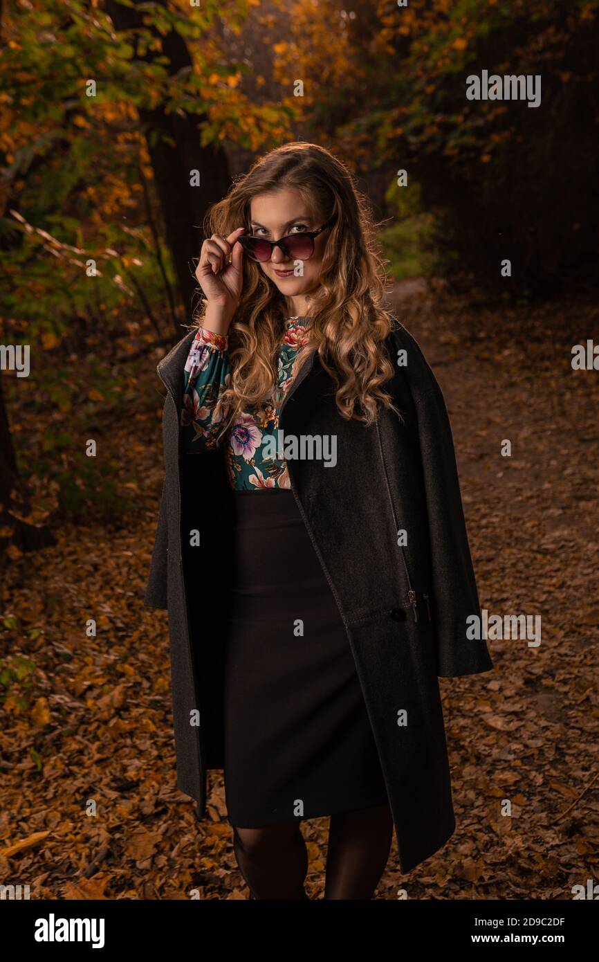 Una chica con gafas de camaleón, se pone de pie y mira hacia abajo en su nariz, sonriendo en un abrigo negro en el otoño en el bosque Foto de stock