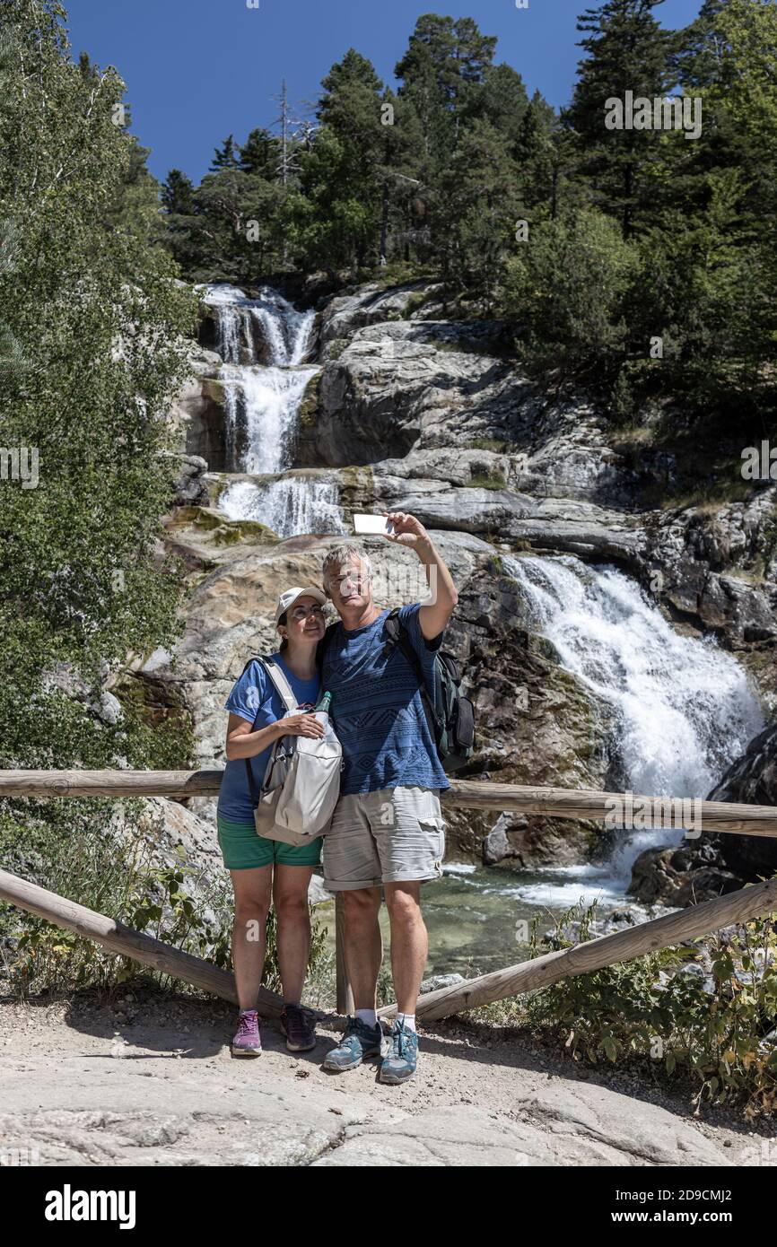 Mochilero de edad media pareja de turistas tomando fotos de sí mismos frente a una cascada en la montaña española. Foto de stock