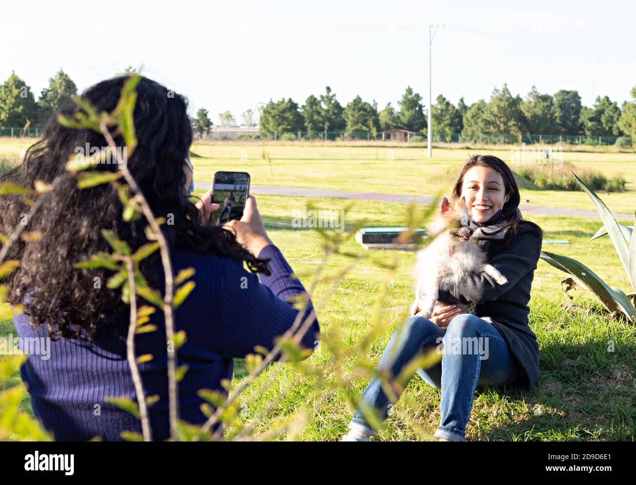 mujer tomando una foto de otra mujer llevando a su perro, mientras se sienta en la hierba en un parque Foto de stock