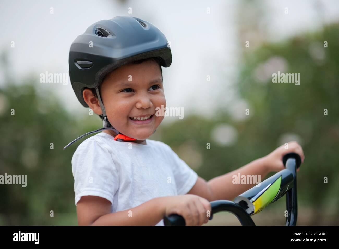 Un niño de cuatro años sonríe, mientras lleva su casco de bicicleta por seguridad y se sostiene en los manillares de la bicicleta. Foto de stock