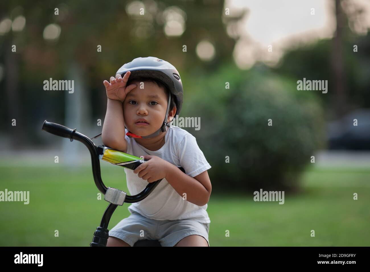 Un niño pequeño que se ve agotado o muestra la dificultad de montar una bicicleta por primera vez al sostener su mano a su frente y parecer infeliz. Foto de stock
