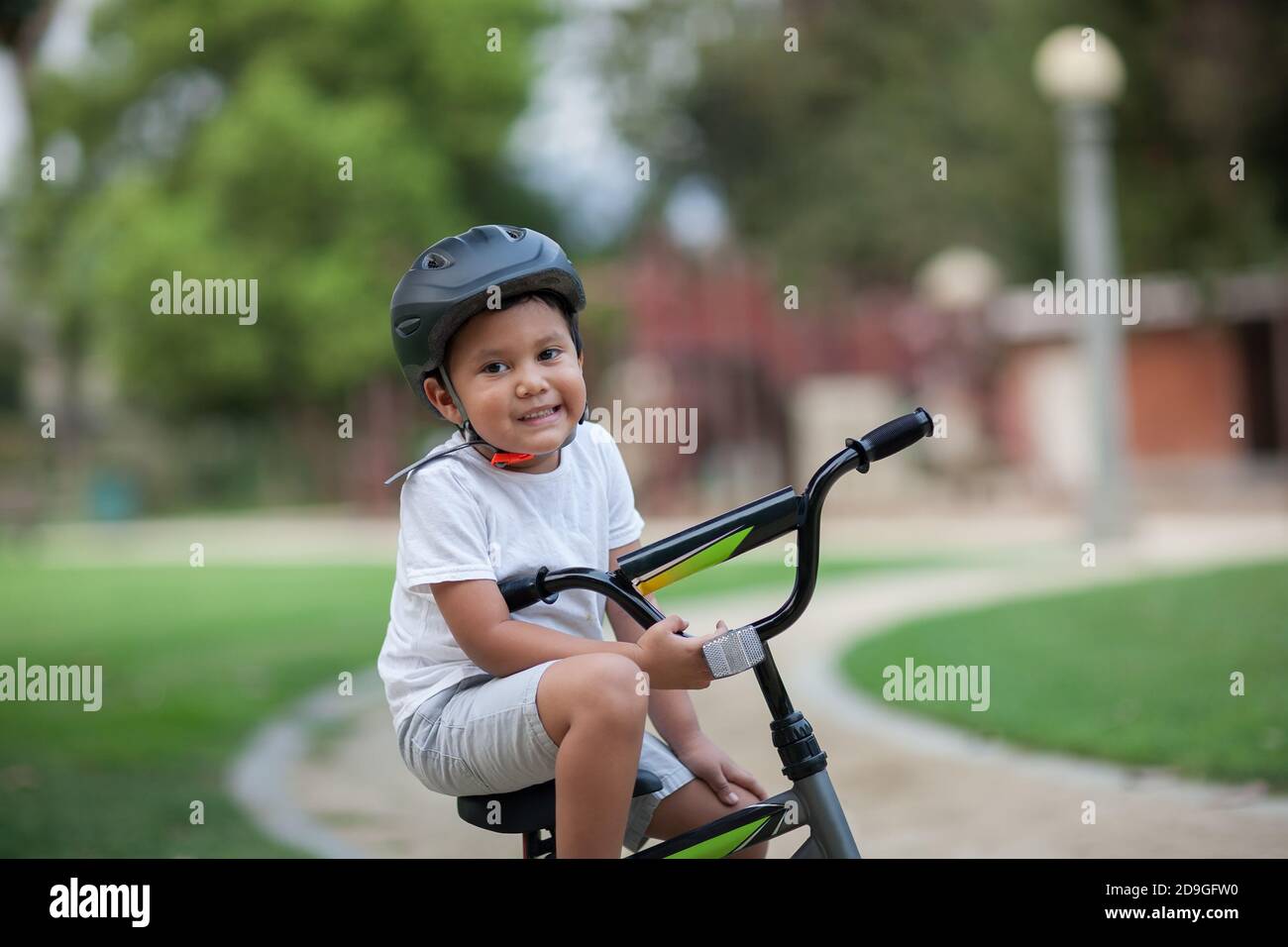 Un niño sano sentado en su bicicleta en un camino del parque que lleva un casco de seguridad, camiseta y pantalones cortos. Foto de stock