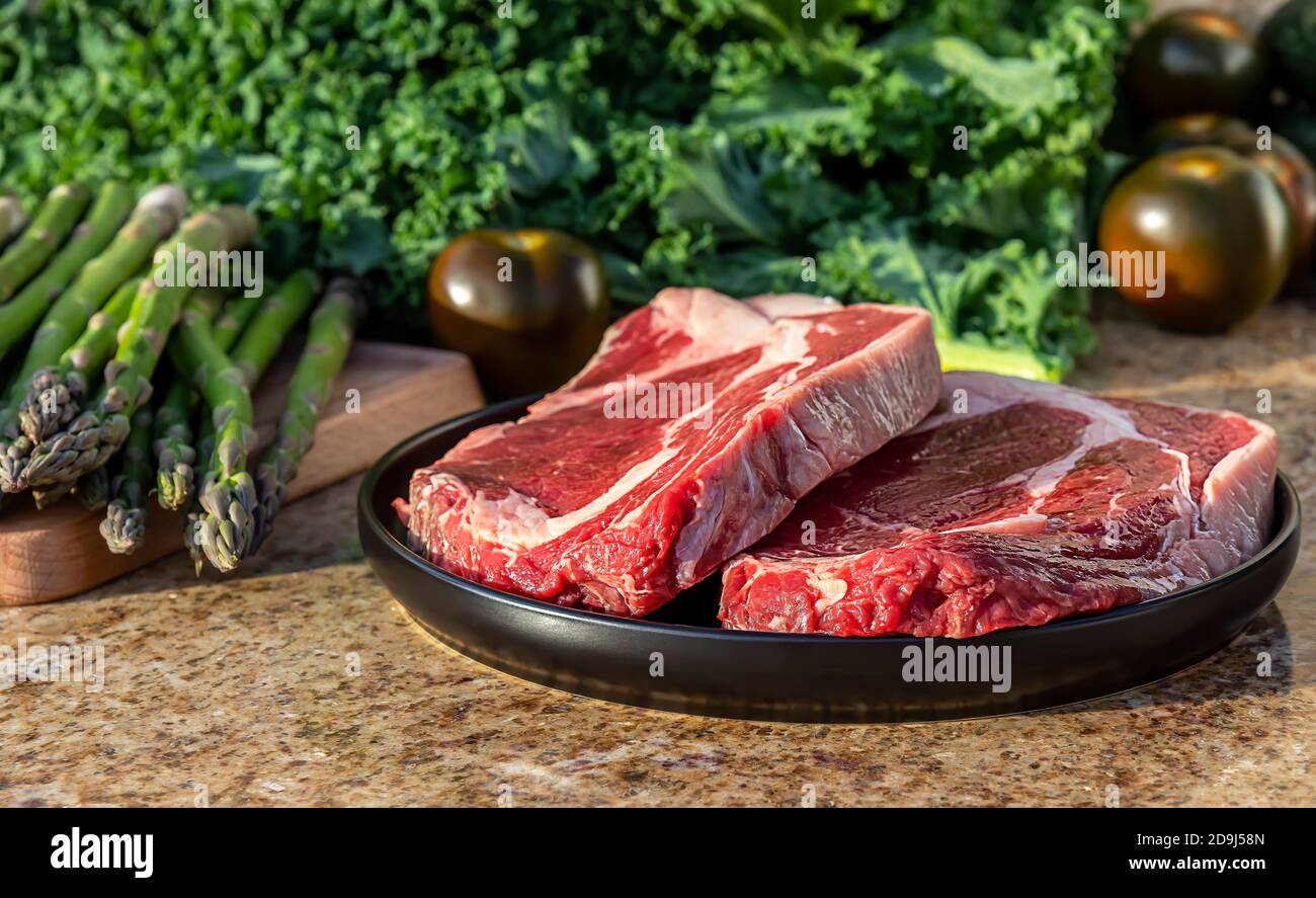Carne cruda, bistec de carne en plato negro con col rizada, espárragos, broccolini y tomates en el fondo. Foto de stock