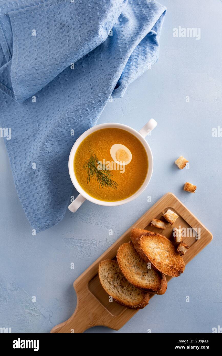 Bouillon en un tazón blanco redondo para sopa, huevo de codorniz y hoja de  hinojo sobre fondo azul. Ropa de cama textil y pan tostado a un lado. Lay  plano. Orientación vertical