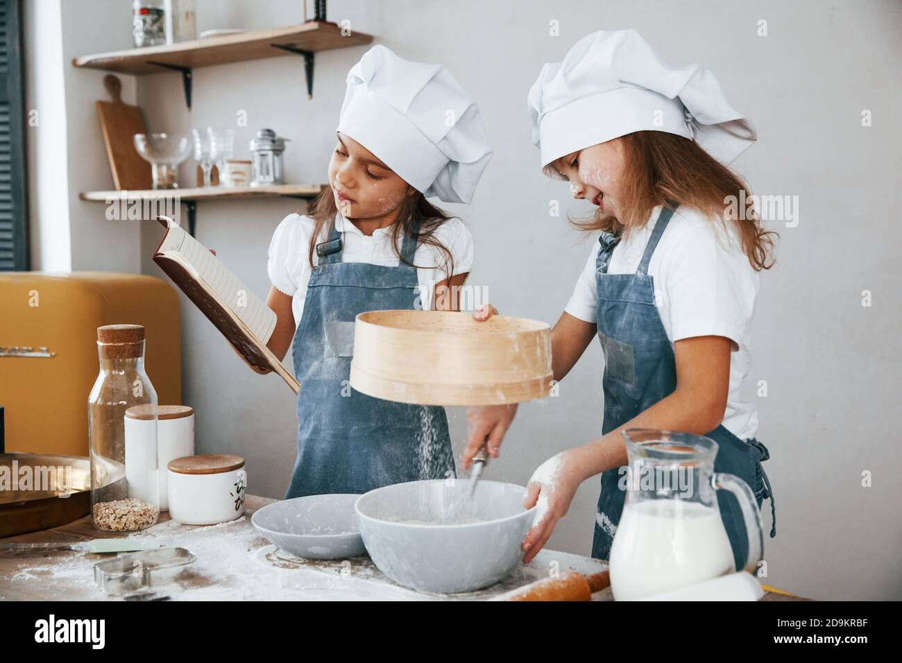Dos niñas en uniforme azul chef preparando la comida usando un tamiz en la cocina y leyendo el libro de recibos Foto de stock