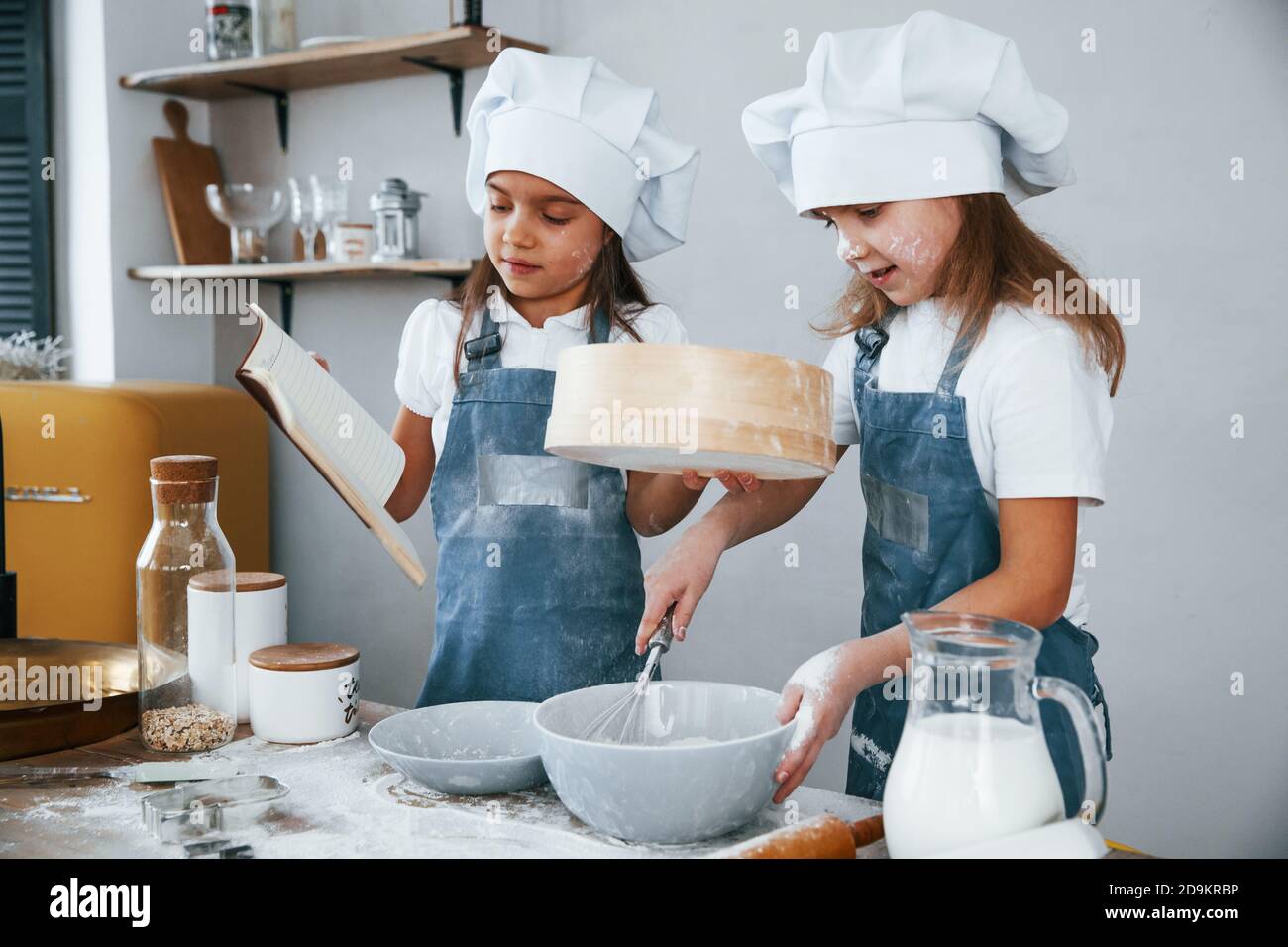 Dos niñas en uniforme azul chef preparando la comida usando un tamiz en la cocina y leyendo el libro de recibos Foto de stock