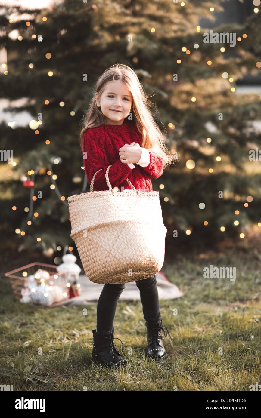 feliz niña de 3-4 años de edad con Jersey de punto rojo sosteniendo bolsa de paja decorar árbol de Navidad con xmas decoración al libre de cerca. Mirando vino Fotografía