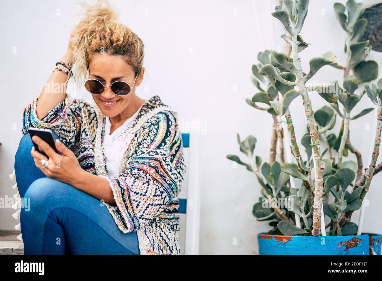 Adulto bonita dama sentarse al aire libre con fondo blanco una planta verde  leyendo el teléfono con una sonrisa - gente feliz en ropa de moda,  accesorios y gafas de sol Fotografía