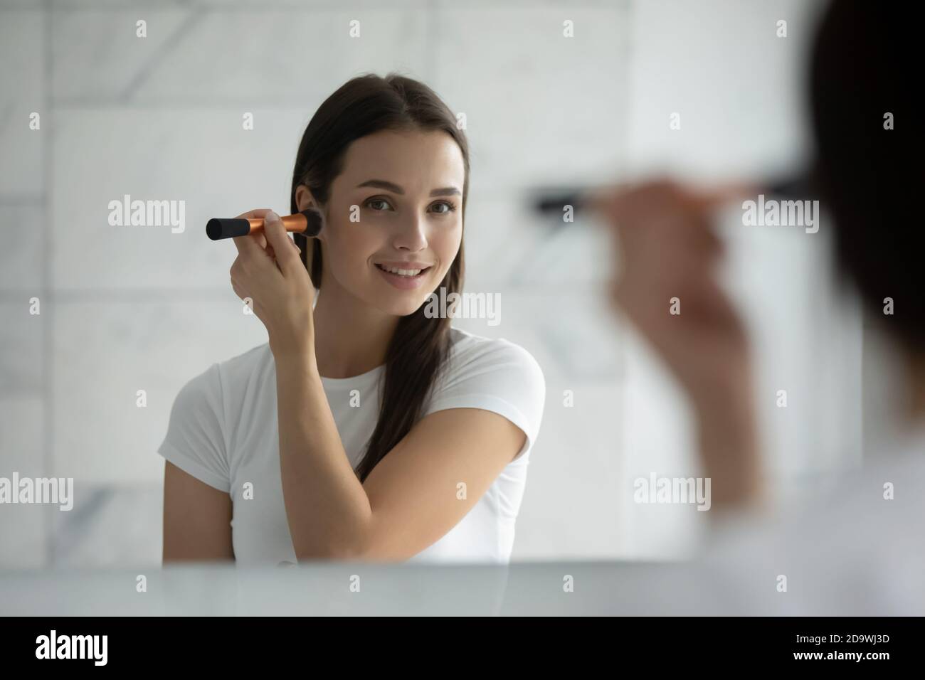 Sonriente joven dama de pie en el espejo aplicando harina en pómulos Foto de stock
