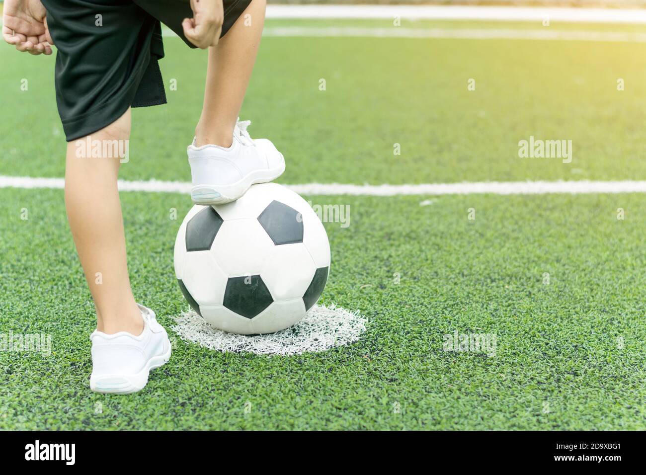 Pies de un niño con zapatillas blancas pisando una pelota de fútbol en el medio del campo de fútbol. Foto de stock