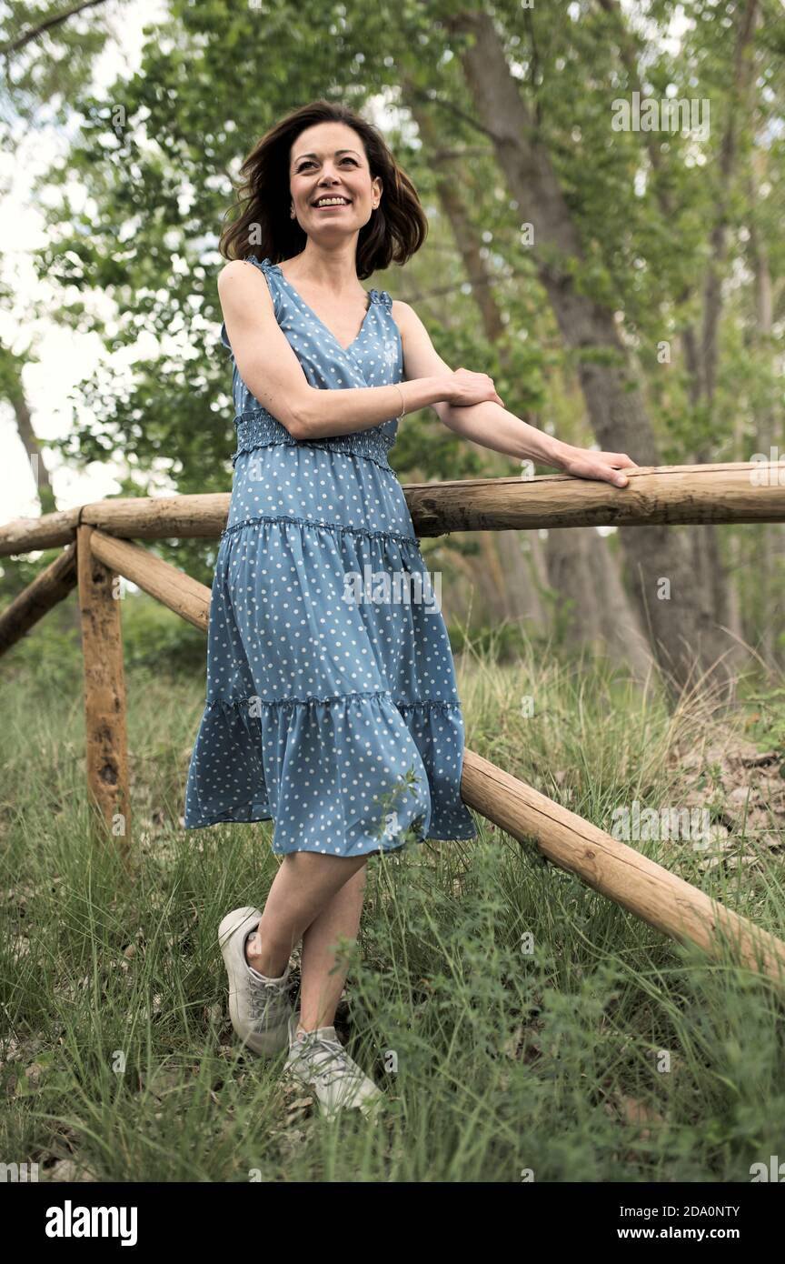 Joven alegre mujer esbelta en vestido mirando fuera mientras está de pie  cerca de una simple valla de madera a lo largo de un estrecho sendero entre  árboles verdes en el bosque