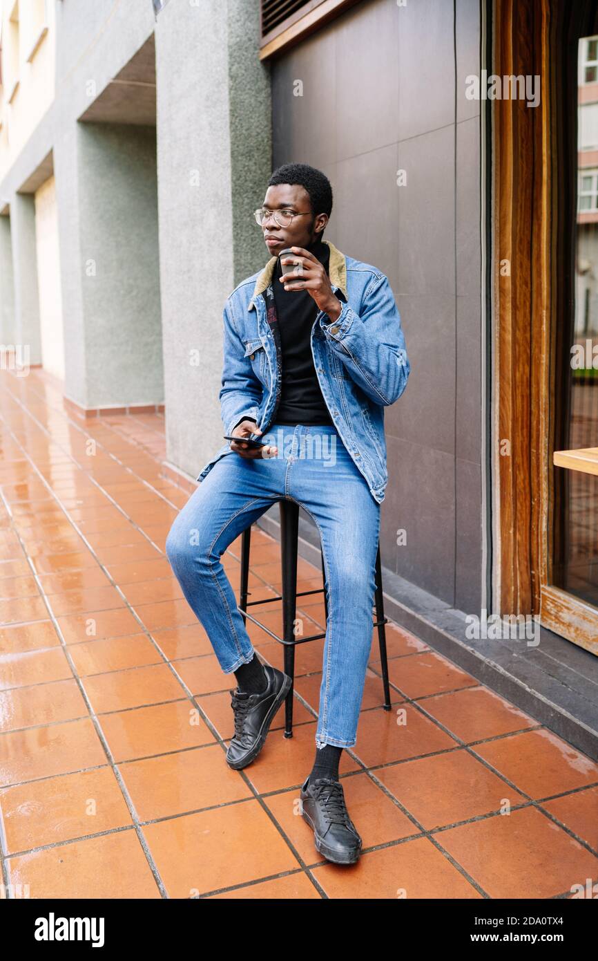 Un hombre afro-americano con ropa vaquera sentado en un taburete en la  calle y beber delicioso café para ir mientras mira lejos Fotografía de  stock - Alamy