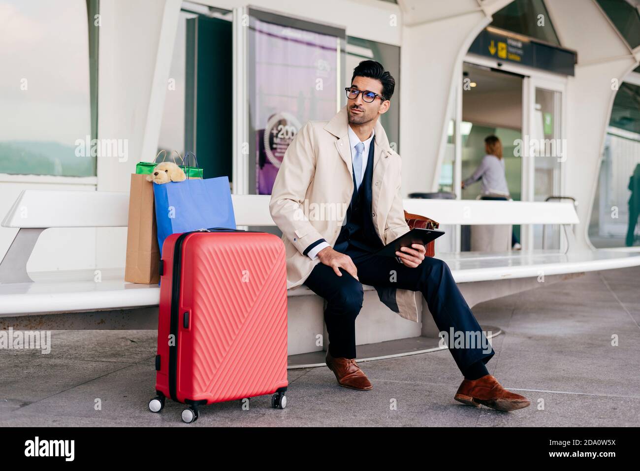 Hombre seguro con peinado en elegante ropa formal con maleta y bolsas de  compras en un banco cerca del aeropuerto y uso tableta Fotografía de stock  - Alamy