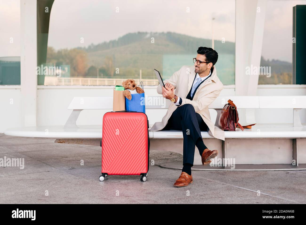 Hombre seguro con peinado en elegante ropa formal con maleta y bolsas de  compras en un banco cerca del aeropuerto y uso tableta Fotografía de stock  - Alamy