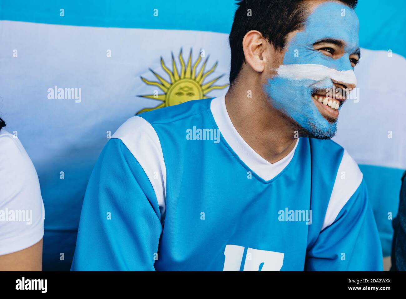 Hombre sentado en el estadio con su cara pintada en colores de bandera Argentina. Devoto fan con la bandera Argentina detrás en el estadio de fútbol. Foto de stock