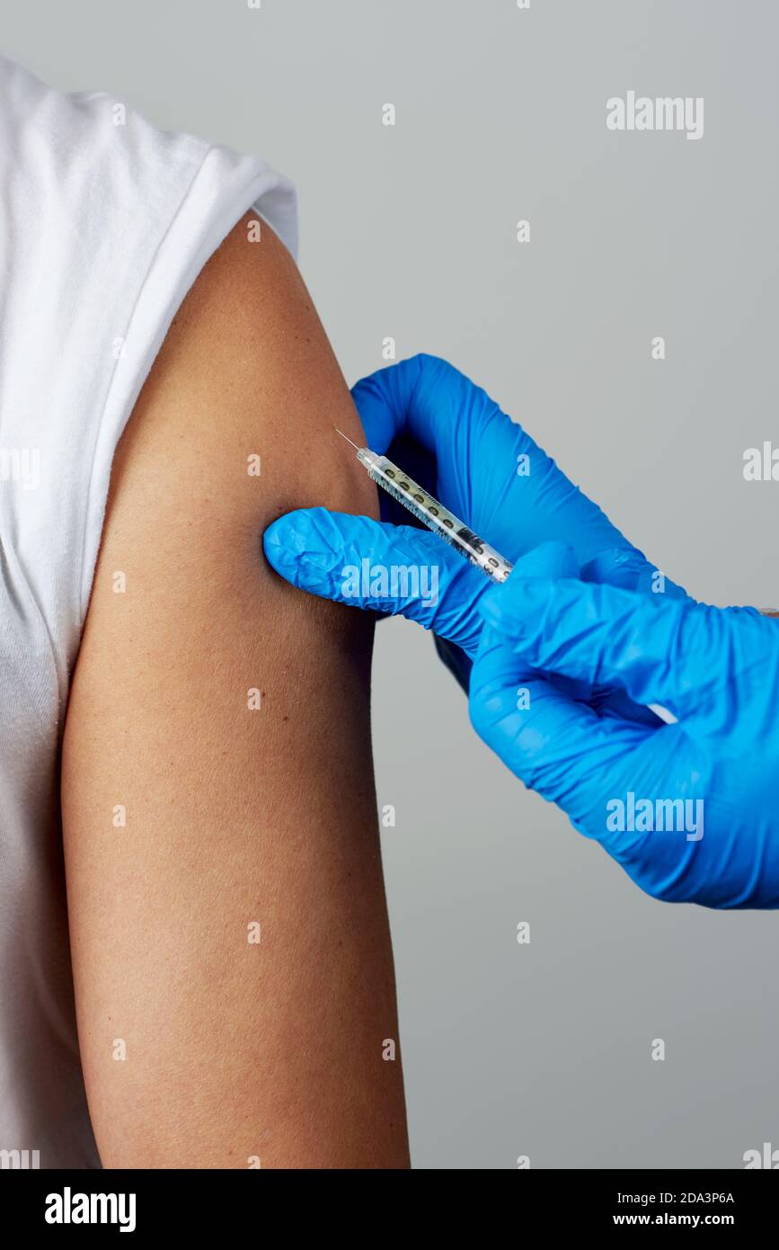 cierre de un médico, con un abrigo blanco y guantes quirúrgicos azules, vacunando a un joven caucásico Foto de stock