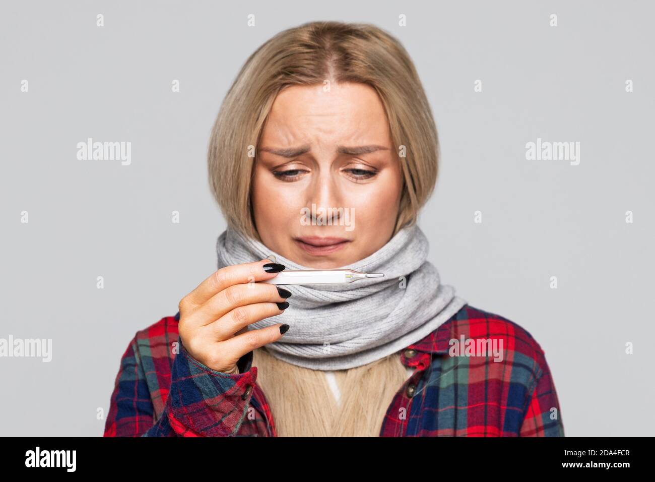 Una mujer malsana se resfrío y mirando el termómetro. Mujer disgustada en la bufanda, camisa de cuadros sintiéndose mal. Frío, enfermedad. Foto de stock