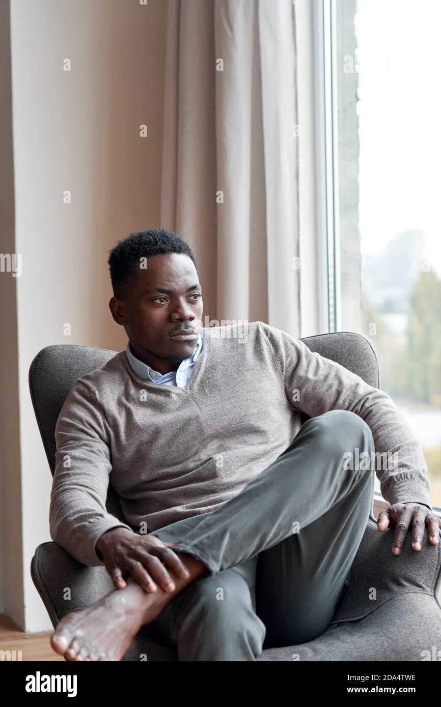 Hombre negro confiado y con estilo sentado en una silla en casa mirando a través de la ventana. Foto de stock