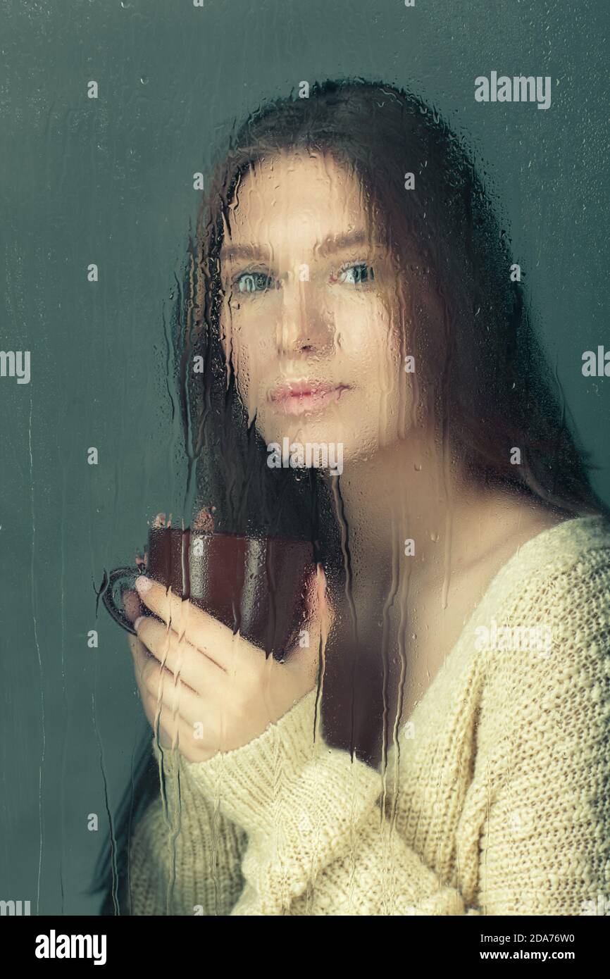 Retrato de una joven rubia con taza de café en las manos. Mujer en lana Jersey de punto detrás de la ventana de vidrio con gotas de lluvia. Bloqueo de auto-aislamiento Foto de stock