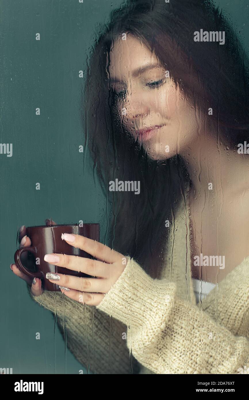 Retrato de una joven rubia con taza de café en las manos. Mujer en lana Jersey de punto detrás de la ventana de vidrio con gotas de lluvia. Bloqueo de auto-aislamiento Foto de stock