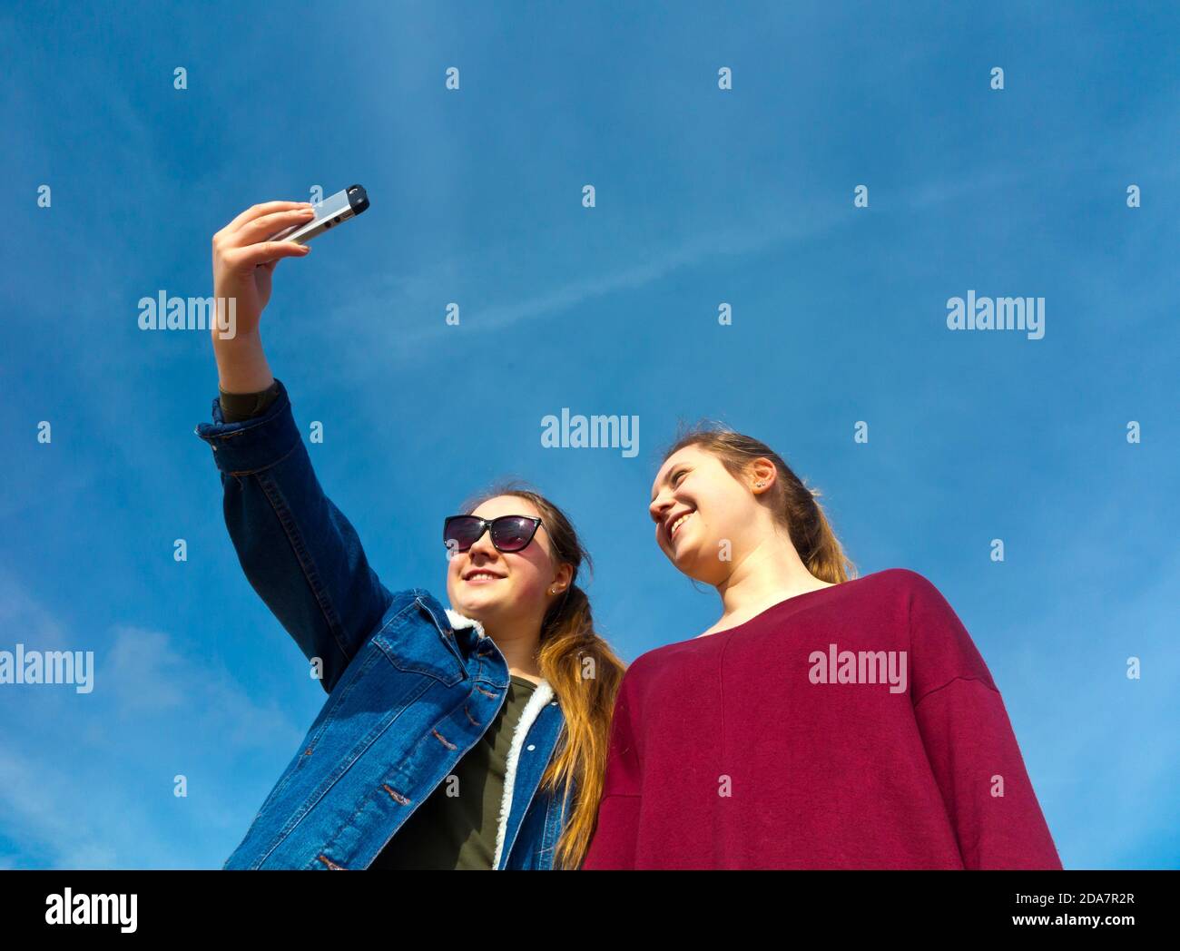 Dos chicas adolescentes que usan un teléfono móvil para tomar fotografías selfie una de la otra con el cielo azul detrás. Foto de stock