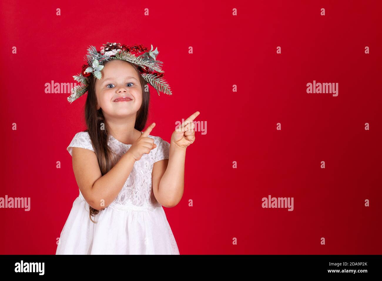 Primer plano de niña sonriente en una corona de Navidad apuntando con sus dedos índice en el espacio vacío para el texto fondo rojo Foto de stock