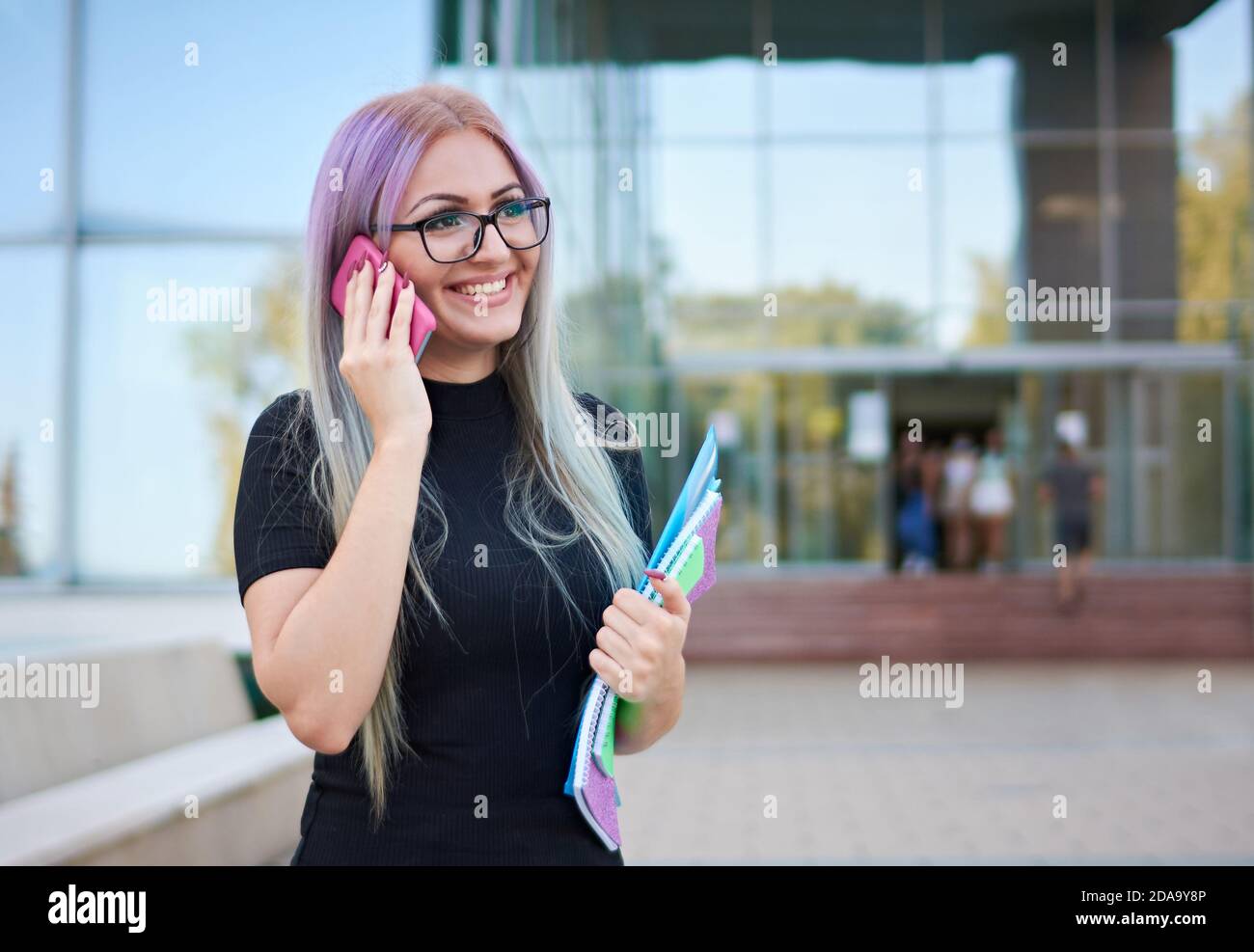 Sonriente niña estudiante de la generación z de pie en un campus en frente de un edificio de la universidad fachada y hablar en el smartphone Foto de stock