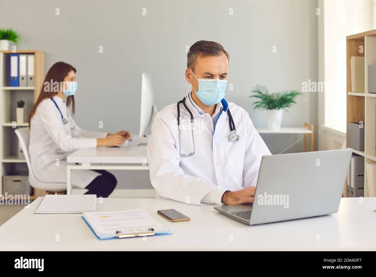Hombre y mujer trabajadores del hospital en máscaras protectoras que trabajan en el consultorio manteniendo la distancia. Foto de stock