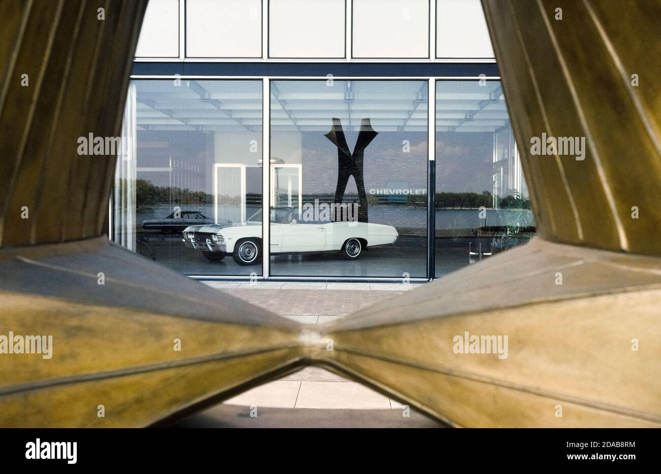 Un Chevrolet Impala convertible de 1967 está enmarcado a través de una escultura al aire libre reflejada en una ventana de exposición en el Centro Técnico General Motors en el suburbio de Detroit Warren, Michigan, EE.UU. Diseñado por arquitectos de padre e hijo de renombre internacional, Eliel y Eero Saarinen, el Centro Tecnológico modernista de GM representó a la América industrial cuando abrió sus puertas en 1956 con 25 edificios repartidos en 320 acres (130 hectáreas). El complejo ampliamente elogiado introdujo un nuevo y novedoso tema arquitectónico: El campus corporativo. Foto de stock