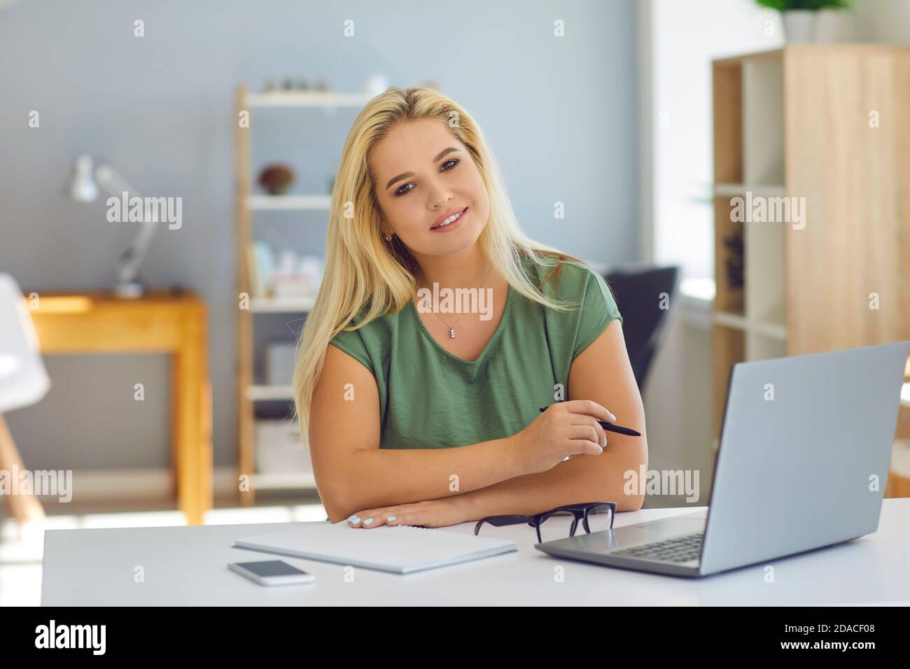 Mujer sonriente trabajando en línea con el portátil y mirando la cámara en casa o en una oficina moderna Foto de stock
