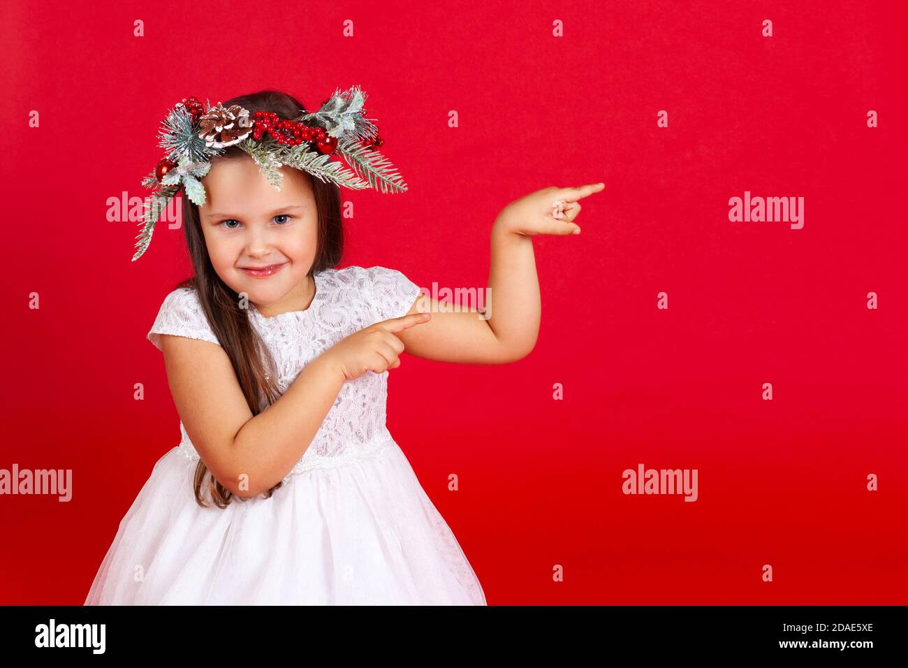 Retrato simulado de una chica sonriente en una corona de Navidad, apuntando con sus dedos índice, sobre un fondo rojo Foto de stock