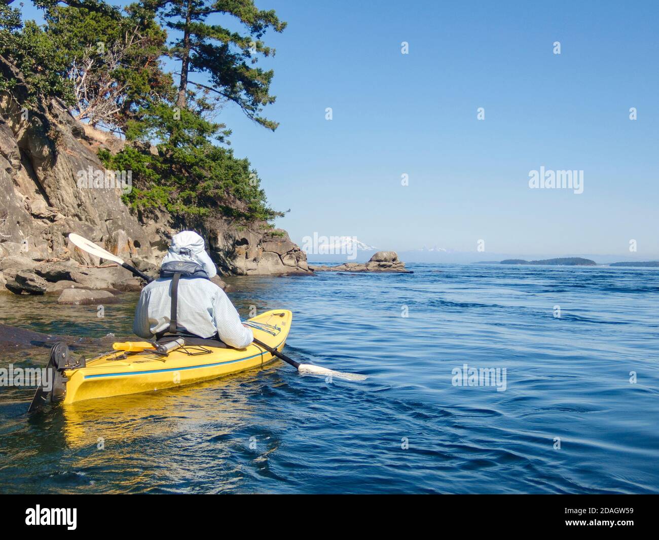 Un hombre, vestido para protegerse del sol de verano, rema un kayak amarillo en las islas del sur del Golfo de Columbia Británica, con Mt. El pico nevado de Baker en la distancia. Foto de stock