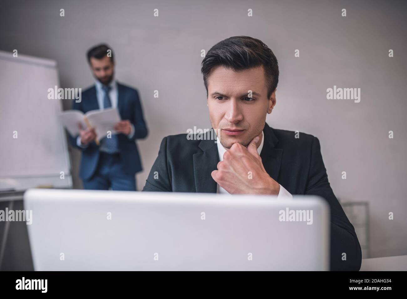 Hombre atento con portátil y colega con papeles detrás Foto de stock