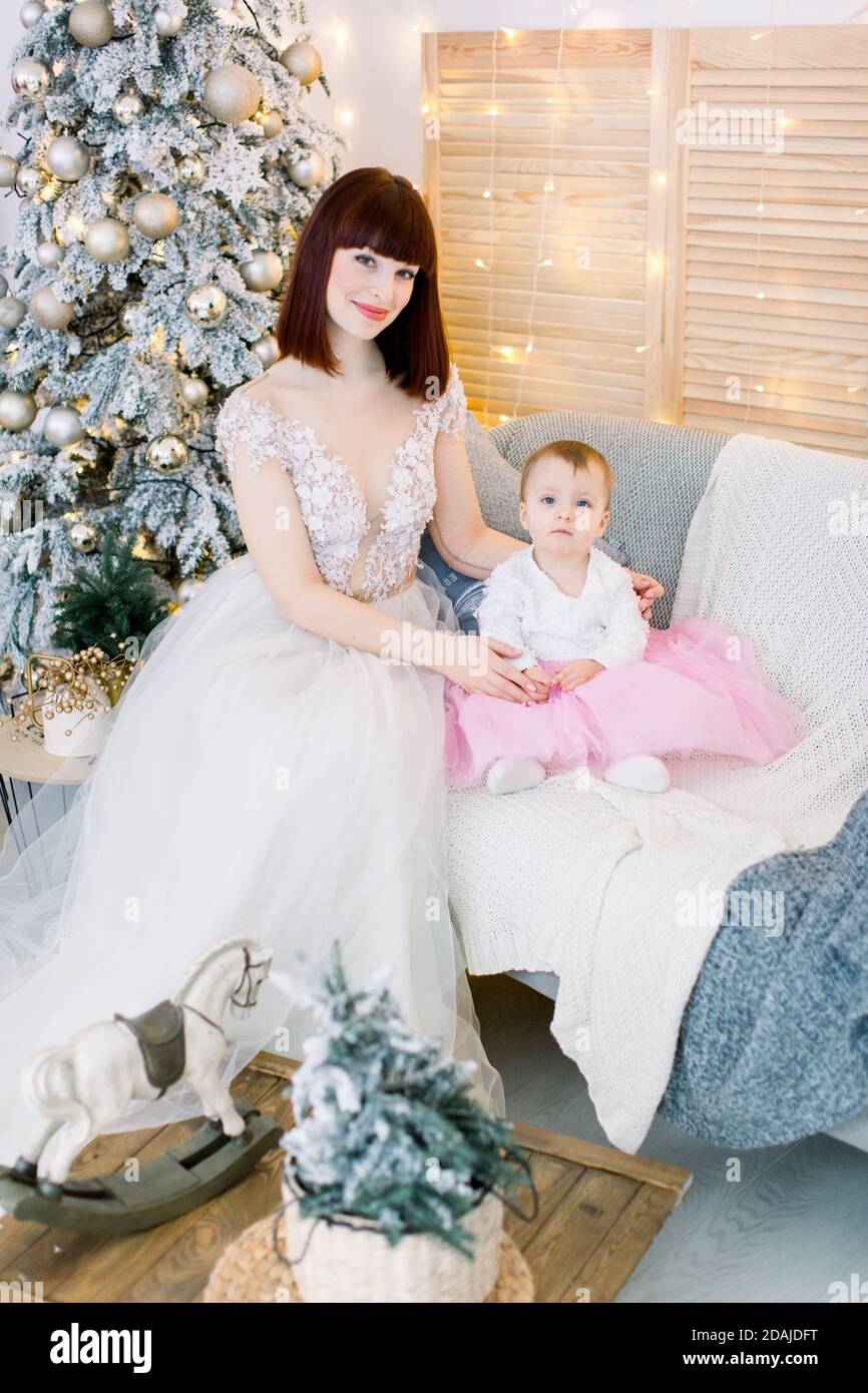 Bonita madre con su bebé niña, vestida con elegantes vestidos de moda,  sentada en un elegante sofá gris sobre el fondo del árbol de Navidad, luces  y Fotografía de stock - Alamy