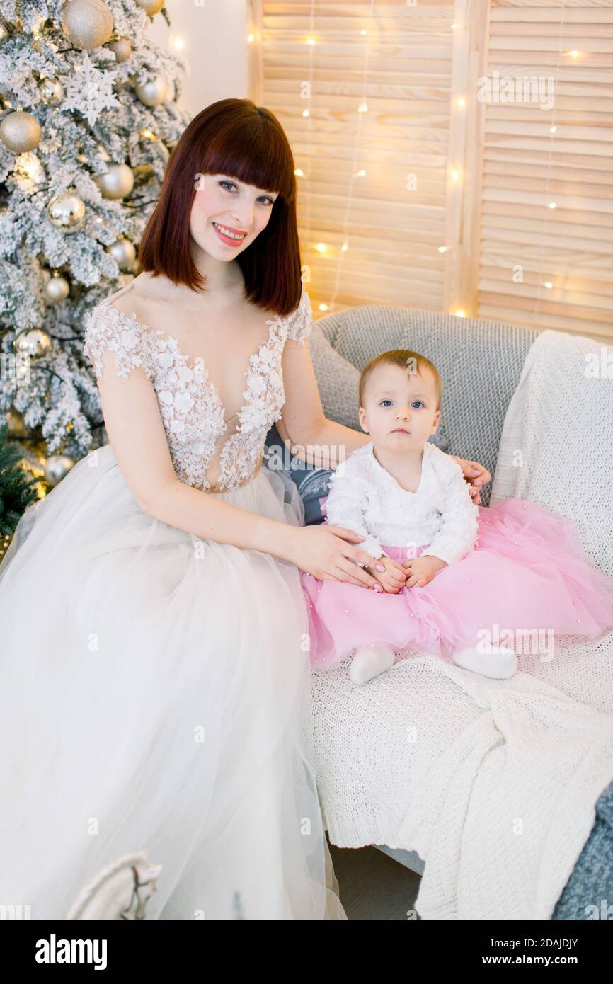 Bonita madre con su bebé niña, vestida con elegantes vestidos de moda, sentada en un elegante sofá gris sobre el fondo del árbol de Navidad, luces y Fotografía stock - Alamy