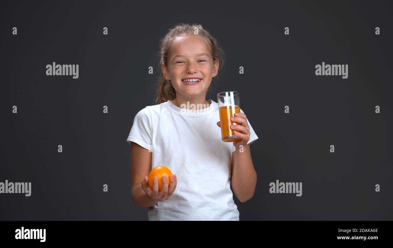 La adolescente disfruta de un vaso de jugo de naranja sosteniendo naranja fresco en otra mano. Concepto de comida saludable. Aislado sobre fondo gris. Espacio de copia Foto de stock