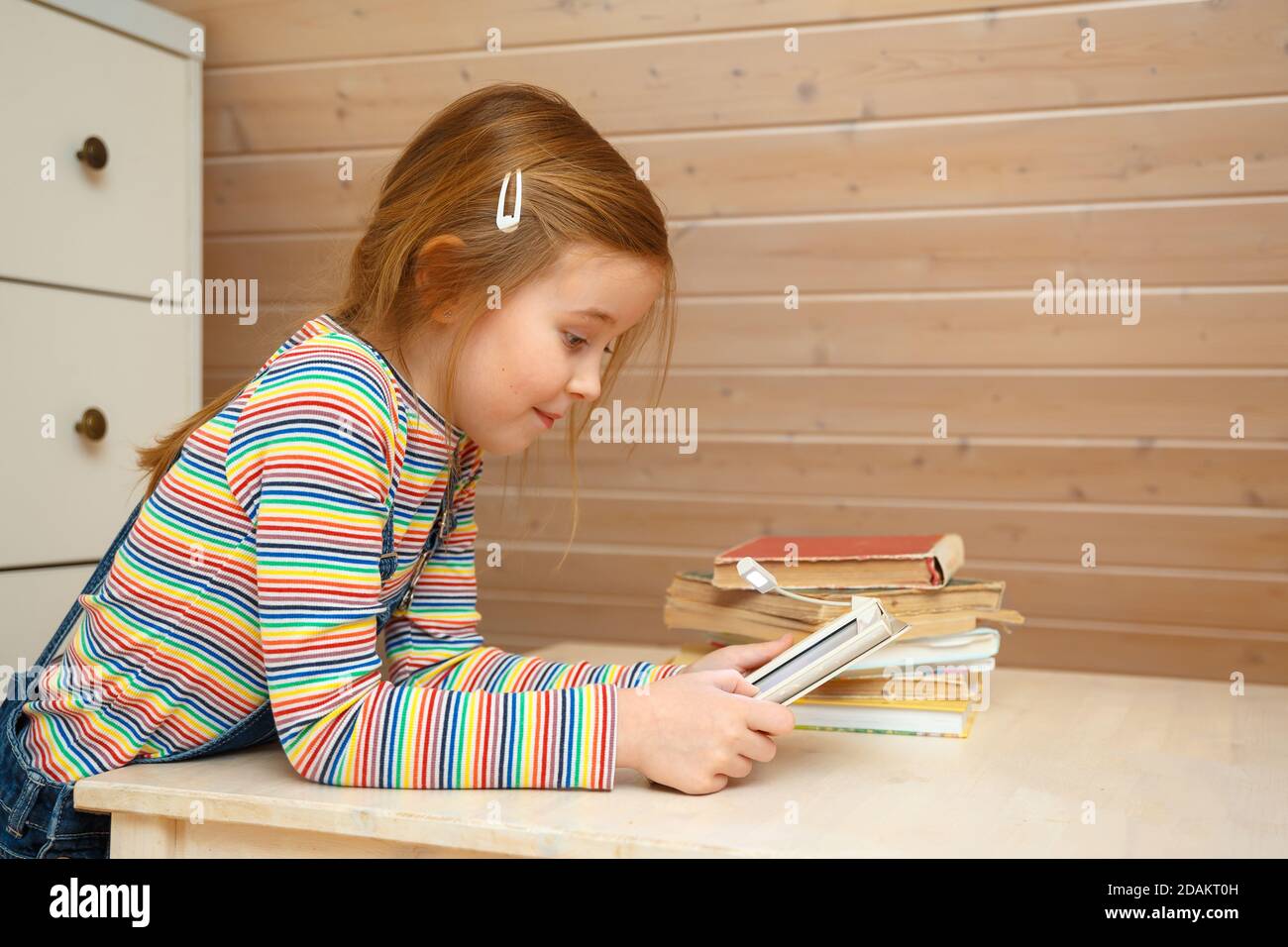 Una niña se sienta en una mesa y lee un libro electrónico. Foto de stock