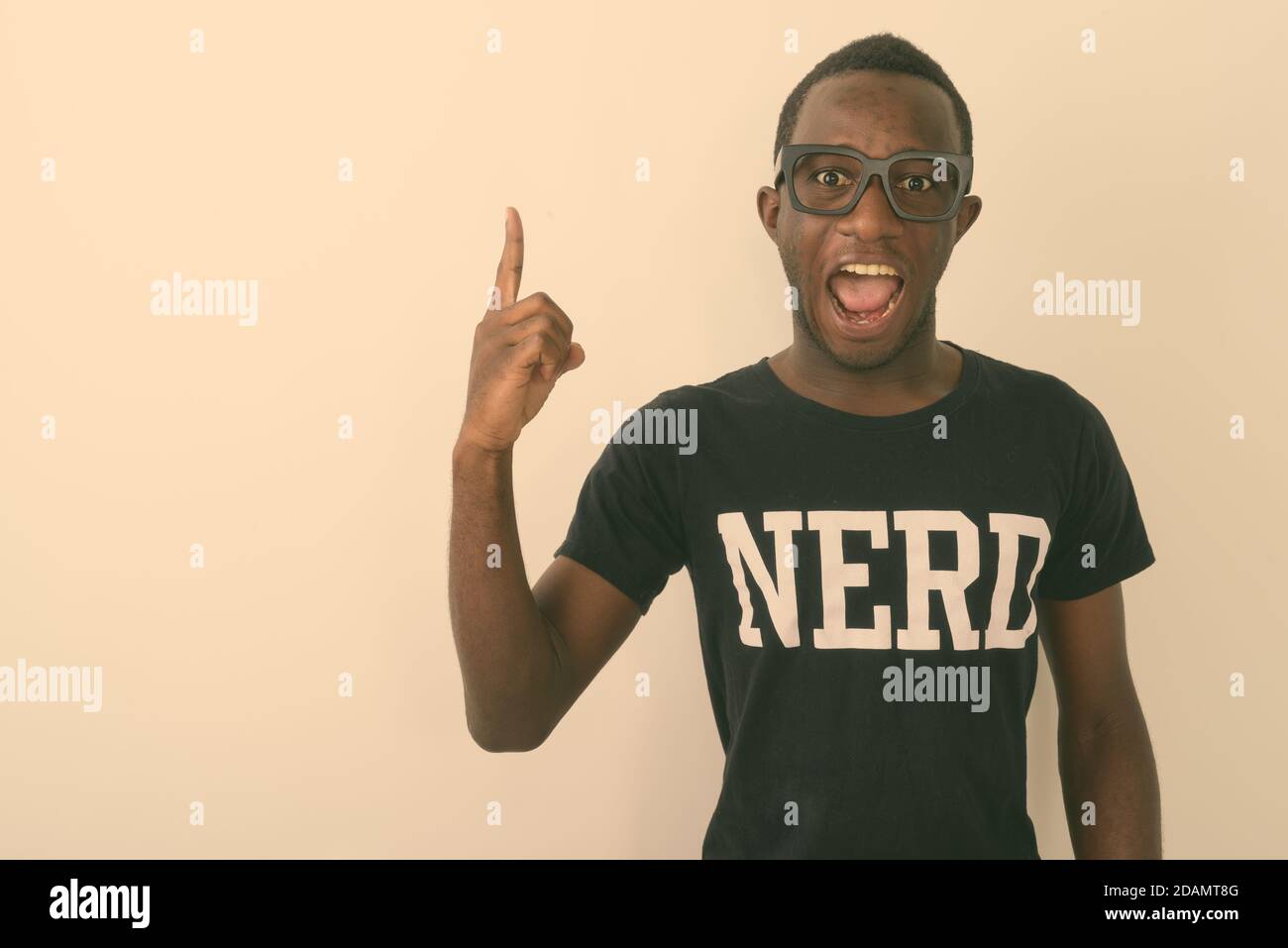 Foto de estudio de joven feliz negro africano geek hombre con Gran idea sonriendo y apuntando con el dedo hacia arriba mientras llevas Nerd camisa y mirando la cámara Foto de stock
